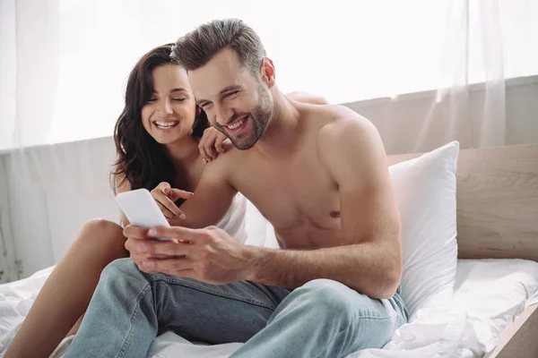 Красивый и улыбающийся мужчина со смартфоном и женщиной, указывающей пальцем на него — стоковое фото