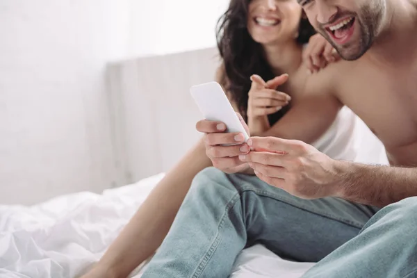 Обрезанный вид улыбающегося мужчины, держащего смартфон, и женщины, указывающей пальцем на него — стоковое фото