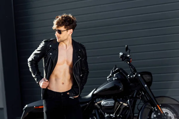 Hombre joven con torso muscular en chaqueta de cuero de pie cerca de la motocicleta negro - foto de stock