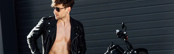Панорамный снимок молодого человека с мускулистым туловищем в кожаной куртке, стоящего рядом с черным мотоциклом — стоковое фото