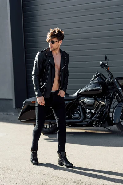 Young man in sunglasses and leather jacket walking near black motorcycle — Stock Photo