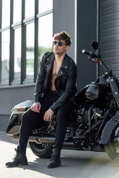 Hombre en chaqueta de cuero negro sentado en motocicleta y mirando hacia otro lado - foto de stock