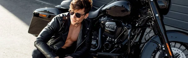 Panoramic shot of young man in glasses and leather jacket sitting near motorcycle — Stock Photo