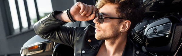 Panoramic shot of young man sitting near motorcycle and holding sunglasses while looking away — Stock Photo