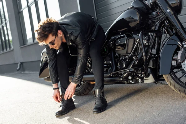 Hombre joven con chaqueta de cuero atando botas mientras está sentado en motocicleta negro - foto de stock