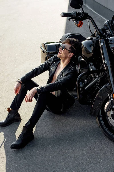 Young man sitting on ground near motorcycle, smiling and looking away — Stock Photo