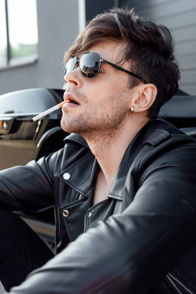 Joven guapo con cigarrillo y gafas de sol sentado cerca de la motocicleta - foto de stock