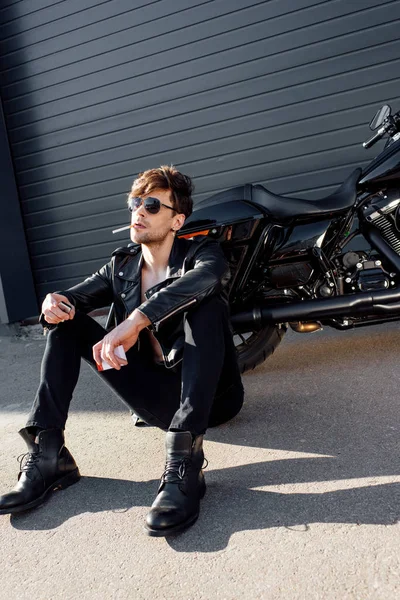 Young man with cigarette sitting on ground near black motorcycle and holding lighter — Stock Photo