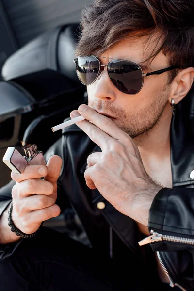 Portraitaufnahme eines jungen Mannes mit Sonnenbrille, der sich Zigarette anzündet, während er in der Nähe eines schwarzen neuen Motorrads sitzt — Stockfoto