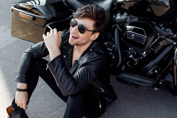 Young man in leather jacket sitting on ground near black motorcycle, smiling and smoking cigarette — Stock Photo