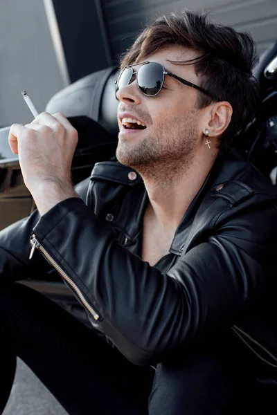 Handsome motorcyclist in sunglasses and leather jacket smoking cigarette while sitting near motorcycle — Stock Photo