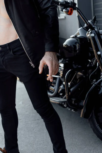 Vista recortada del hombre con el torso desnudo sosteniendo el cigarrillo y de pie no lejos de la motocicleta - foto de stock