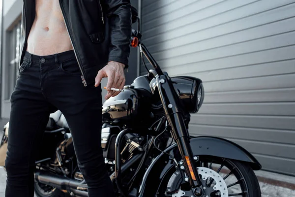 Vista recortada del hombre con el torso desnudo sosteniendo cigarrillo y de pie cerca de la motocicleta - foto de stock