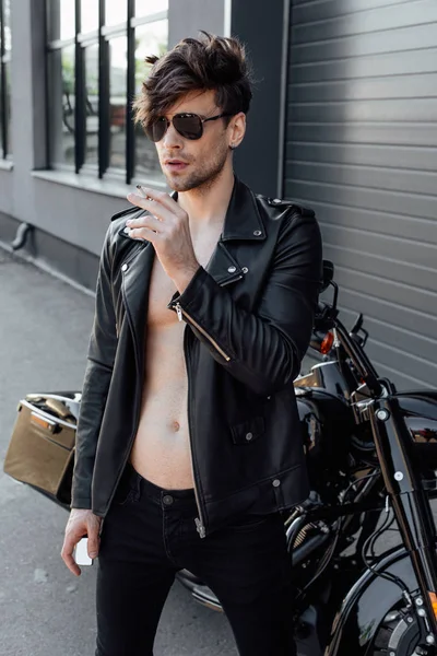 Young motorcyclist with muscular naked torso standing near black motorcycle smoking and holding lighter — Stock Photo