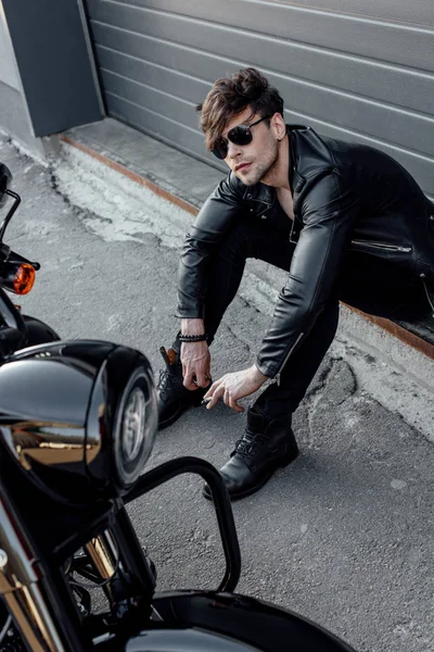 Schöner Motorradfahrer mit Sonnenbrille sitzt auf dem Boden neben Motorrad — Stockfoto