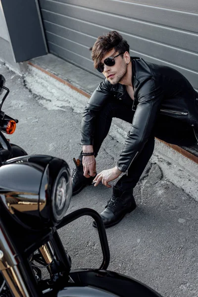 Guapo motociclista en gafas de sol sentado en el suelo cerca de la motocicleta - foto de stock