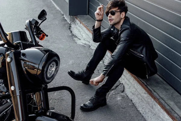 Guapo motociclista en gafas de sol sentado en el suelo cerca de la motocicleta y fumar cigarrillo - foto de stock