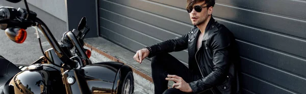 Panoramic shot of handsome motorcyclist in sunglasses sitting on ground near motorcycle and holding cigarette — Stock Photo