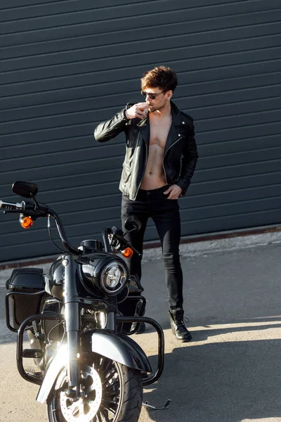 Full length view of motorcyclist with muscular torso standing near motorcycle and holding cigarette — Stock Photo