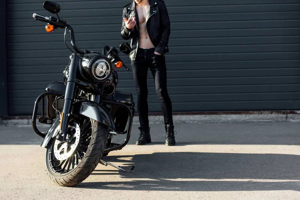 Обрезанный вид мотоциклиста, курящего и стоящего рядом с черным мотоциклом — стоковое фото