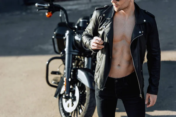 Cropped view of man with muscular torso in leather jacket looking away and smoking cigarette — Stock Photo