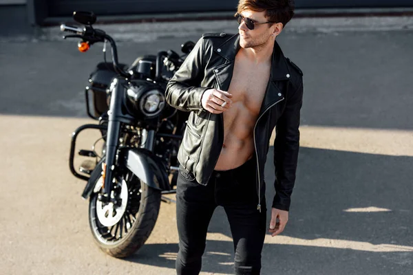 Full length view of handsome young man with muscular torso in leather jacket looking away and smoking cigarette — Stock Photo