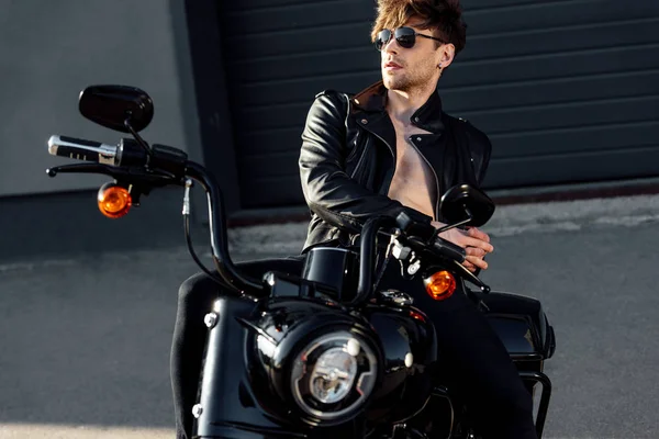 Handsome young motorcyclist with naked in sunglasses and leather jacked torso leaning on black motorcycle — Stock Photo