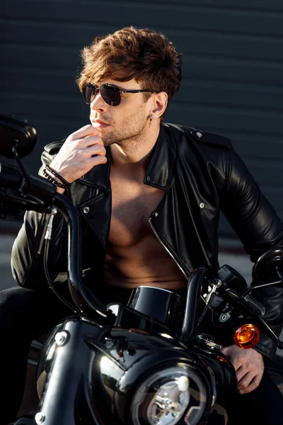 Молодой человек в солнцезащитных очках и черной кожаной куртке с обнаженным туловищем смотрит в сторону и сидит на мотоцикле — стоковое фото