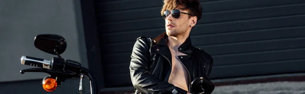 Panoramic shot of handsome young motorcyclist with naked in sunglasses and leather jacked torso leaning on black motorcycle — Stock Photo