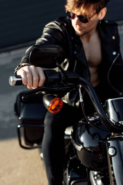 Foco seletivo do jovem sentado na motocicleta à luz do sol, segurando alça e olhando para longe — Fotografia de Stock