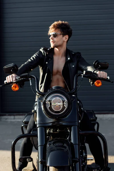 Guapo motociclista con torso desnudo muscular en chaqueta de cuero sentado en motocicleta y agarraderas - foto de stock