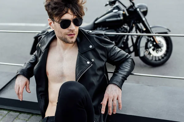 Selective focus of man with naked torso in leather jacket sitting on ground with motorcycle on background — Stock Photo
