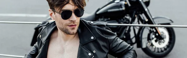Panoramic shot of man with naked torso in leather jacket sitting on ground with motorcycle on background — Stock Photo