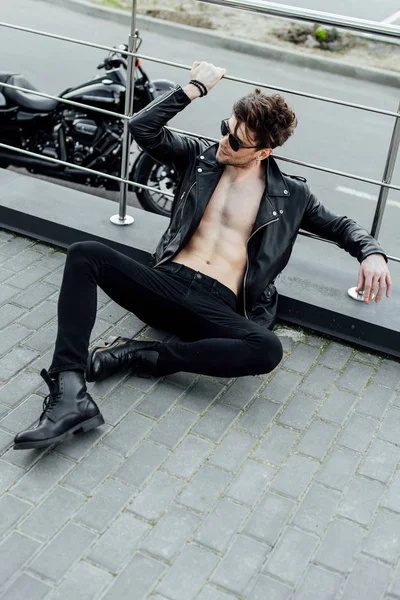 Sexy motorcyclist in leather jacket with naked torso sitting on ground and holding metal fence — Stock Photo