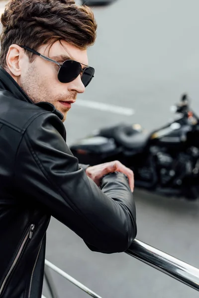 Selective focus of young man in black leather jacket and sunglasses standing near metal fence with motorcycle on background — Stock Photo