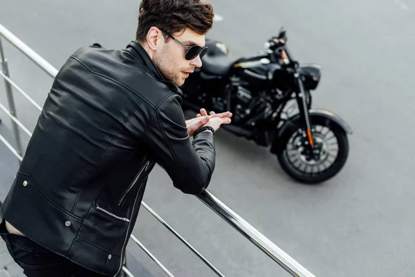 Jeune homme en cuir noir veste debout près de la clôture métallique avec moto sur fond — Photo de stock