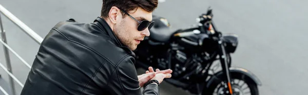Panoramic shot of young man in black leather jacket standing near metal fence with motorcycle on background — Stock Photo