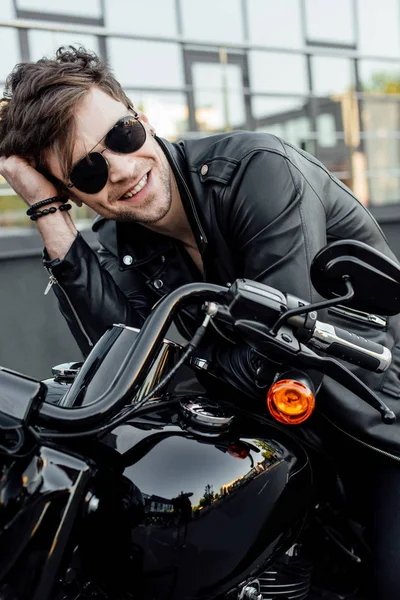 Guapo joven motociclista en gafas de sol sosteniendo la cabeza con la mano, sonriendo y mirando hacia otro lado mientras está sentado en la motocicleta - foto de stock