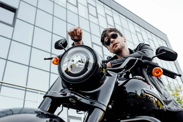 Low angle view of handsome young motorcyclist in sunglasses looking away while sitting on motorcycle — Stock Photo