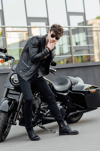 Schöner junger Motorradfahrer steht neben Motorrad, schaut weg und berührt Kinn — Stockfoto