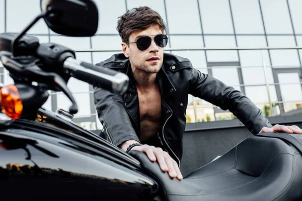 Selective focus of handsome man in sunglasses and black leather jacket leaning on motorcycle and looking forward — Stock Photo