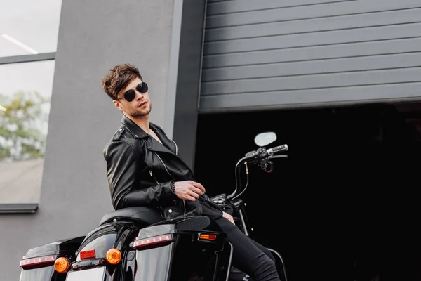 Motociclista em óculos de sol e jaqueta de couro sentado na motocicleta perto da garagem aberta e olhando para a câmera — Fotografia de Stock