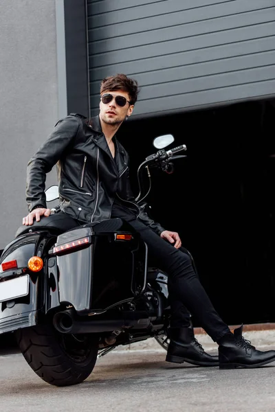 Full length view of motorcyclist in sunglasses and leather jacket sitting on motorcycle near opened garage — Stock Photo