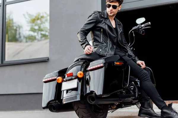 Handsome motorcyclist in sunglasses and leather jacket sitting on motorcycle near opened garage — Stock Photo