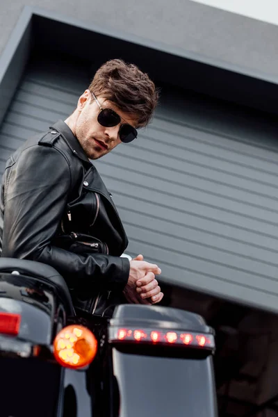 Stylish young man in sunglasses leaning on motorcycle and looking at camera — Stock Photo