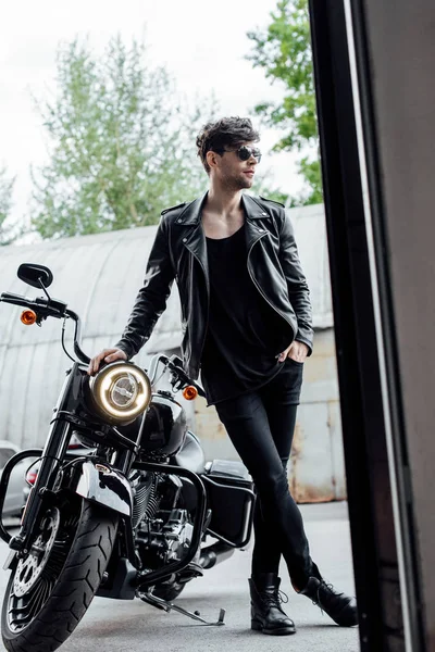 Vista completa de hombre guapo en chaqueta de cuero de pie cerca de la motocicleta y apoyándose en ella - foto de stock