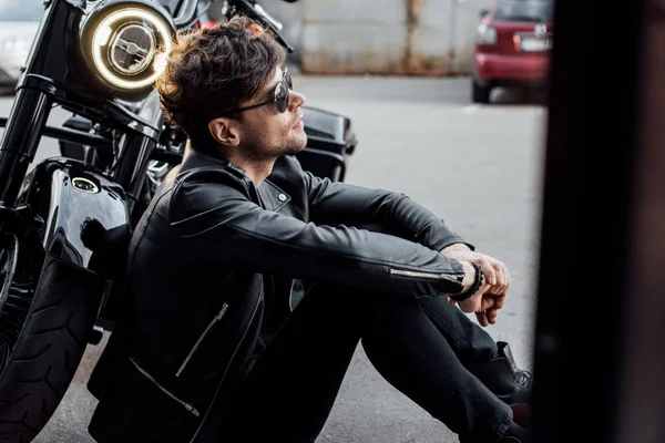 Side view of handsome motorcyclist sitting on ground near motorcycle with lamp lighting up — Stock Photo