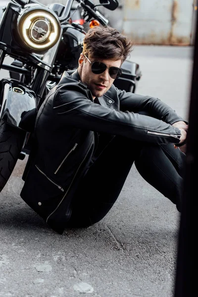 Joven guapo sentado en asfalto cerca de la motocicleta - foto de stock