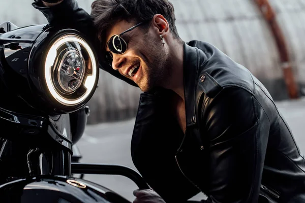 Joven guapo apoyado en la motocicleta y sonriendo mientras mira la lámpara - foto de stock