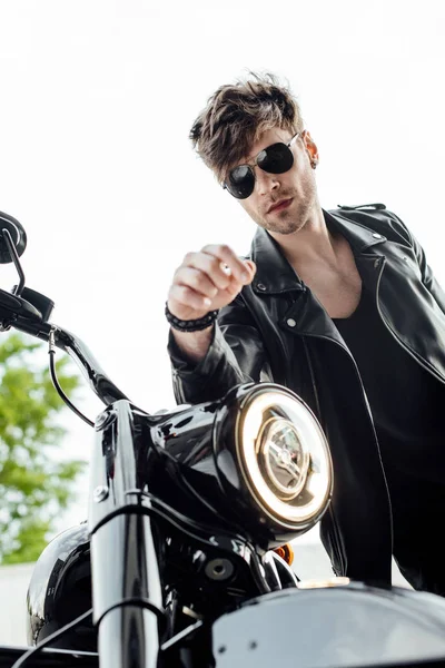 Low angle view of handsome motorcyclist standing near motorcycle and leaning on handlebars — Stock Photo